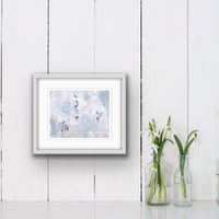 An abstact acrylic painting on paper, double matted and in a white frame.Colours are a soft grey/blue, white and brown.The painting is hanging on a white wall beside two glass frames filled with white flowers.