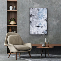 This is a large rectangular abstract painting hanging vertically. It is highly textured in grey/blue, sandy taupe, white and black. It is hanging in a room with a grey wall,over a round brown table and a beige chair, Beside it is a narrow shelf.