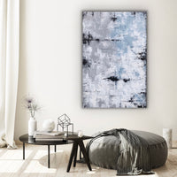 This is a large rectangular abstract painting hanging vertically. It is highly textured in grey/blue, sandy taupe, white and black. It is hanging in a room with a white wall, over a black round table and a grey pouff.  A white curtain is visable from the left.