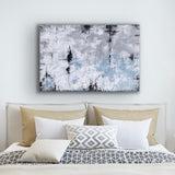 This is a large rectangular abstract painting hanging horizontally. It is highly textured in grey/blue, sandy taupe, white and black.It is hanging over a bed with a beige headboard, tan ,cream and grey patterned cushions are placed on the bed.