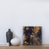 A small square abstract artwork. The colours are brown, black , and gold.  The painting is sitting on a shelf with two vases and some books.