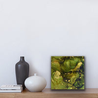 This is a small square abstract artwork on a birch panel. A botanical inspired piece in dark olive green and metallic gold. The painting is sitting on a shelf with two vases and some books.