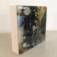 A side view of a small square abstract artwork. The colours are brown, black  and gold. The sides are natural wood.