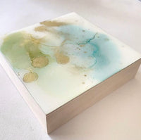 A side view of small square abstract painting in alcohol ink and resin. The background color is white and there is a pattern of soft blue, green and gold in the center. The sides area natural wood colour.