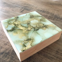 A side view of a small square abstract artwork. The colours are light sage green and gold. The sides are natural wood.