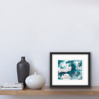An abstract acrylic painting on paper in a black frame. The painting is of a wave, in dark teal and white. There is collaged text in sections of the painting.The painting sits on a shelf beside two vases and some books.