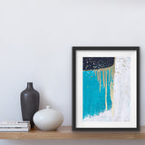 An abstract acrylic painting on paper, in a white mat and framed in a modern black frame. The painting is turquoise, black and white, with accents of metallic gold. There are three main sections, one is turquoise, one is black and one is white. A gold line runs between the shapes and there are some splashes of gold and white at the top, and some gold dripping down from the top.The painting is sitting on a shelf beside two vases ans some books.